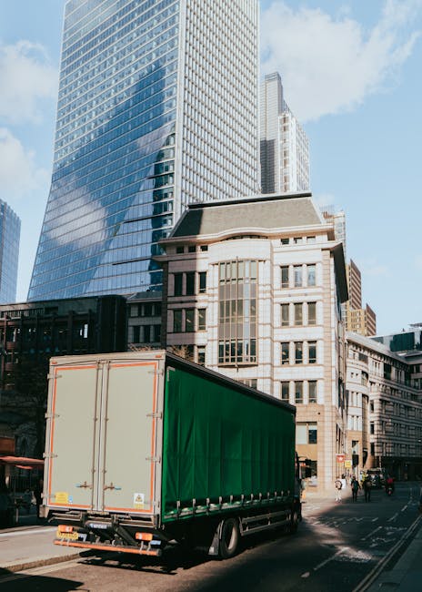 View of a busy urban street lined with multi-storey shopfronts and residential buildings on both sides, with pedestrians walking along the pavement and a cyclist riding in the middle of the road. In t