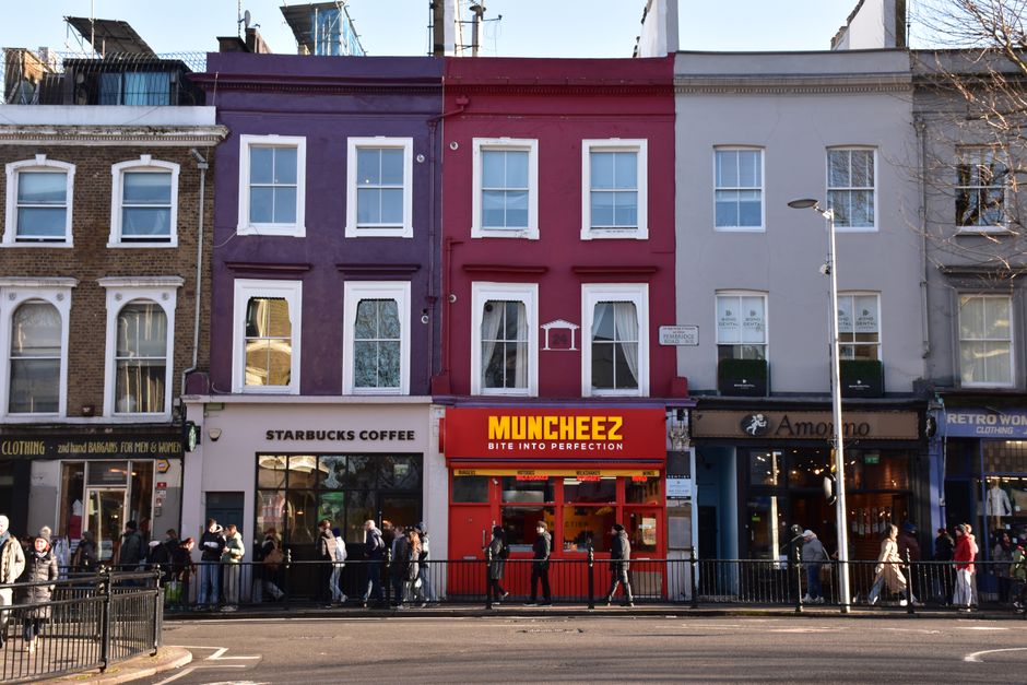 View of a row of shops on a busy urban street in Soho, showing three ground-floor retail stores with signage for Starbucks Coffee, Muncheez, and Amo Amo. The buildings are three stories tall, with the
